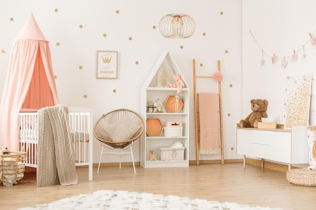 Sillón dorado y escalera en un interior de dormitorio femenino de color pastel con una cuna blanca con dosel