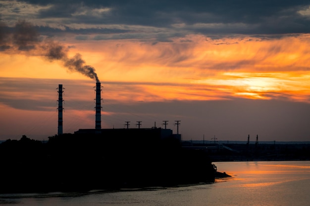 Silueta al atardecer de una planta industrial con una pipa humeante ubicada en un río Contaminación industrial y concepto de calentamiento global