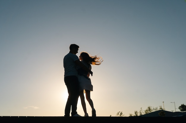 Silueta de amorosa pareja abrazándose al atardecer sobre fondo de cielo despejado en la noche, de cuerpo entero. Feliz pareja caucásica de enamorados con paseo romántico al aire libre disfrutando de tiempo juntos.