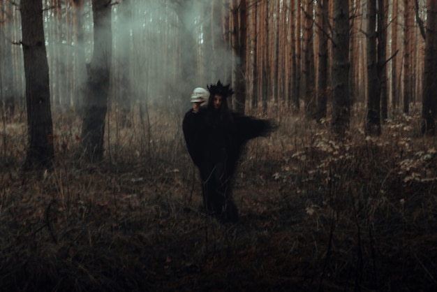 Silueta borrosa de una malvada bruja negra con una calavera en sus manos realizando un ritual satánico oculto en un bosque oscuro y sombrío
