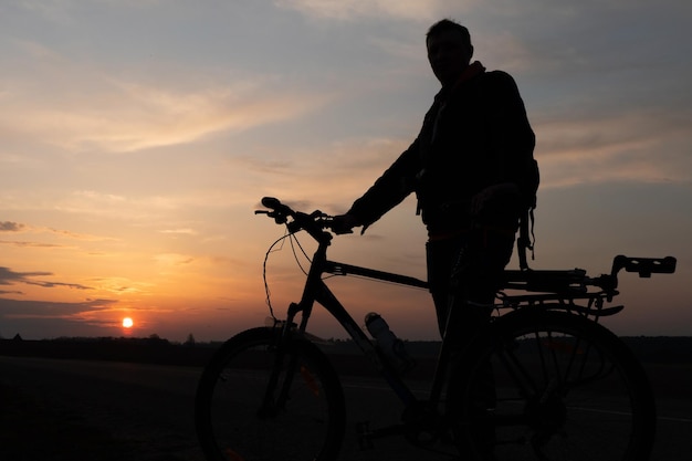 La silueta de un ciclista contra el fondo del sol y el hermoso cielo El contorno de un hombre de pie junto a su bicicleta contra el fondo de la puesta de sol