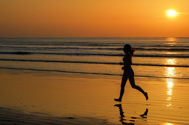 Silueta de corredor de mujer corriendo en la playa al atardecer con concepto de reflexión, fitness y deporte