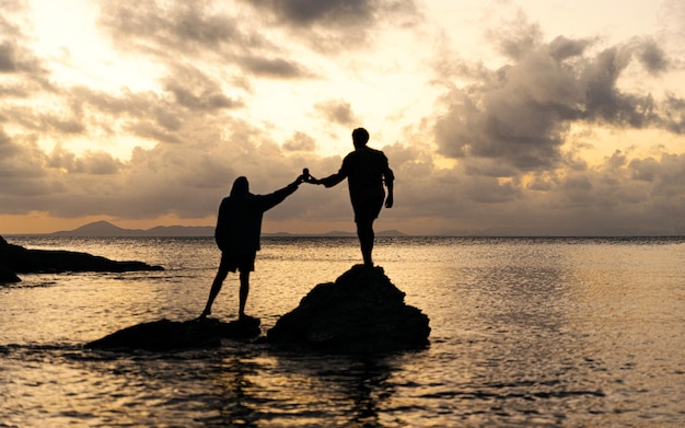 Silueta de dos amigos de pie sobre las rocas en el océano compartiendo una bebida al atardecerCopiar espacio