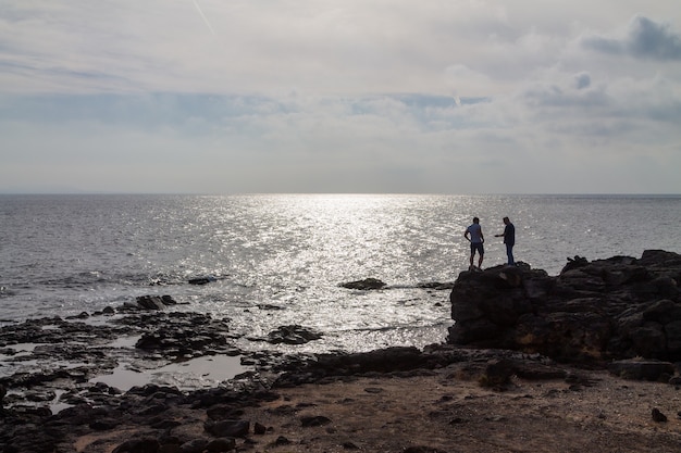 Silueta de dos hombres de pie sobre una gran roca junto al océano. Lanzarote, España.