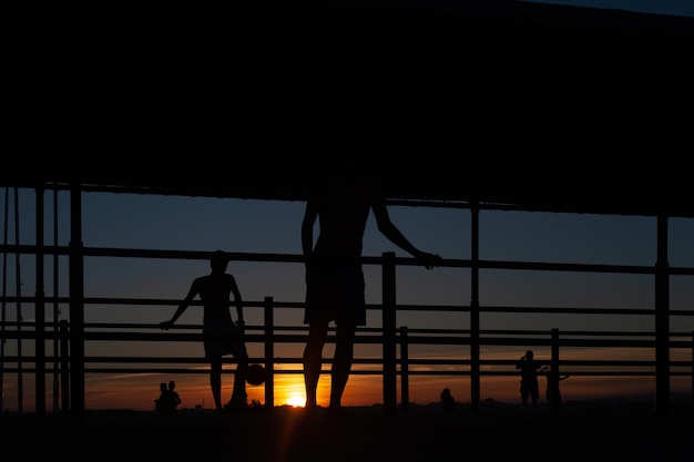 silueta de dos jóvenes jugando al fútbol en una pasarela portuaria en una playa de Portugal al atardecer