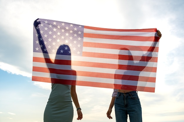 Silueta de dos mujeres jóvenes amigos sosteniendo la bandera nacional de Estados Unidos en sus manos parados juntos. Chicas patrióticas celebrando el día de la independencia de Estados Unidos.