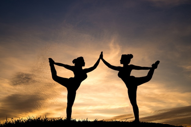 Silueta de dos yoga hermosa de las muchachas que coloca el arco permanente que tira de actitud en el parque en el cielo hermoso de la puesta del sol.