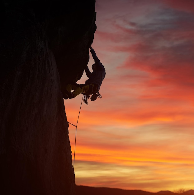 Silueta escalador masculino escalada en roca tirando hacia arriba con ambas manos Puesta de sol cielo naranja sobre fondo Ángulo bajo Copiar espacio