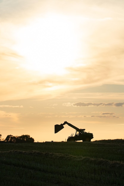 Silueta de una excavadora en funcionamiento con un balde levantado contra el fondo de la puesta de sol y las nubes El campo es operado por maquinaria agrícola La temporada de cosecha o siembra del cultivo