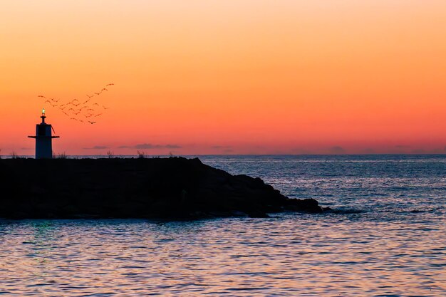 Silueta de un faro en una playa al atardecer. Una bandada de pájaros volando al atardecer. Un hombre solitario.