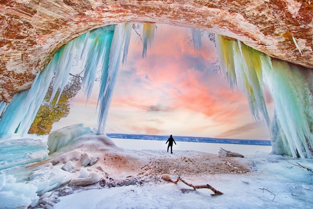 Silueta de figura humana en la entrada a la impresionante cueva de hielo durante el amanecer de invierno con i azul y verde