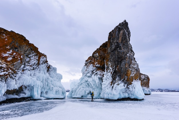 La silueta de un hombre de chaqueta amarilla con mochila se encuentra cerca de dos rocas cubiertas de carámbanos. Lago Baikal en tiempo nublado.