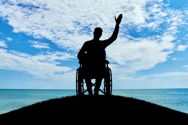 Silueta de un hombre discapacitado en silla de ruedas agitando su mano en la cima de una colina cerca del mar. El concepto de personas con discapacidad