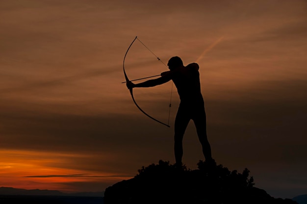 Silueta de un hombre guapo con un arma antigua arco y flecha sobre un fondo de cielo y puesta de sol