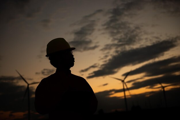 Silueta de hombre ingeniero trabajando y sosteniendo el informe en la estación generadora de energía de la granja de turbinas eólicas en la gente de mountainThailand