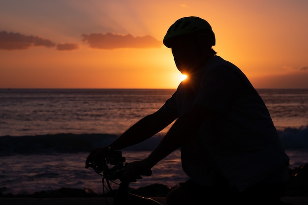 Silueta de hombre maduro en bicicleta en la playa con una magnífica puesta de sol naranja. Rayo de sol en el horizonte