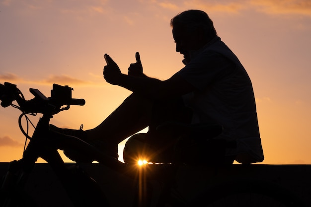 Silueta de hombre maduro sentado cerca de su bicicleta a la luz del atardecer mediante teléfono móvil