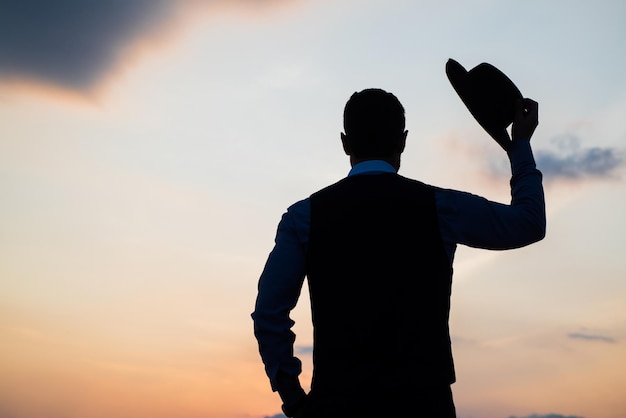 Silueta de hombre de negocios con sombrero contra la soledad del cielo del atardecer