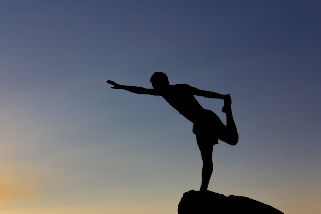 Silueta de hombre practicando yoga al atardecer sobre una roca en la naturaleza. Iluminar desde el fondo. Copie el espacio.