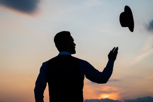 Silueta de hombre subiendo su sombrero en el cielo del amanecer, despreocupado.