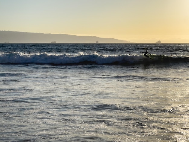 Silueta de hombre surfeando olas al atardecer en la playa con buques de carga por el puerto de Valparaíso en Chile