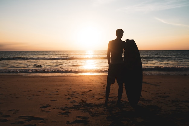 Silueta de hombre surfista llevando sus tablas de surf en la playa al atardecer con luz solar