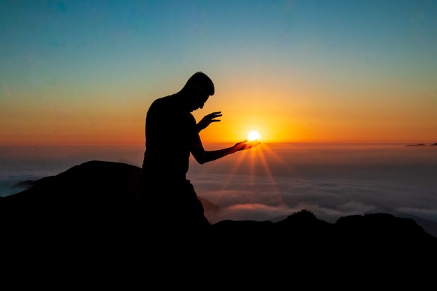 silueta de un hombre tocando el sol con ambas manos en la cima de una montaña