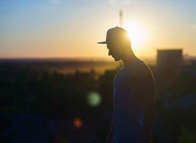 Silueta de hombre vestido con gorra en el fondo del paisaje urbano durante la puesta de sol