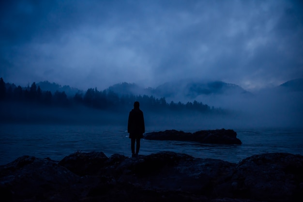 Silueta humana en una espesa niebla azul en el fondo de colinas boscosas y río de montaña. Figura femenina solitaria en una orilla misteriosa. Contemplación, meditación, unidad en la naturaleza.