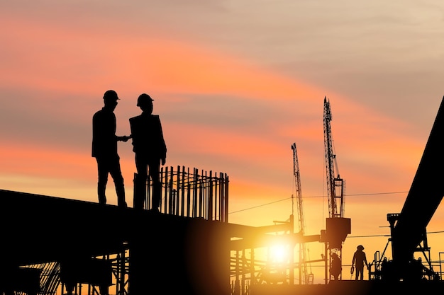 Silueta del ingeniero y el equipo de trabajadores revisando el proyecto en el fondo del sitio de construcción, Construcción de una línea de tren de transporte masivo en progreso con un sitio de infraestructura pesada al atardecer en el tiempo de la tarde