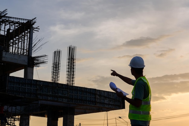 Silueta de un ingeniero masculino de pie en un sitio de construcción al atardecer Ingenieros de construcción inspeccionan la calidad y los planes de diseño de proyectos de construcción industrial