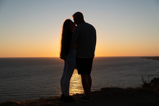 Silueta de una joven pareja romántica disfrutando de una velada en un acantilado sobre el mar con un rojo