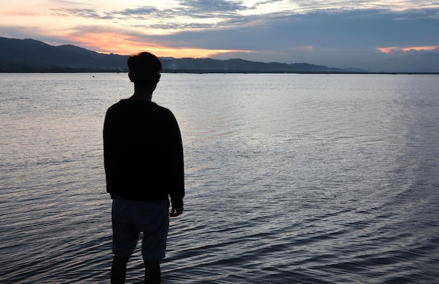 Silueta de un joven de pie junto al lago disfrutando de la puesta de sol. ambiente tranquilo en la naturaleza