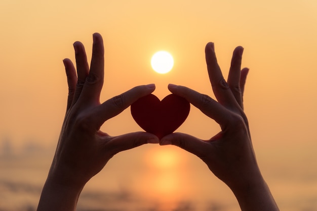 Silueta de mano sosteniendo corazón rojo durante el atardecer de fondo.