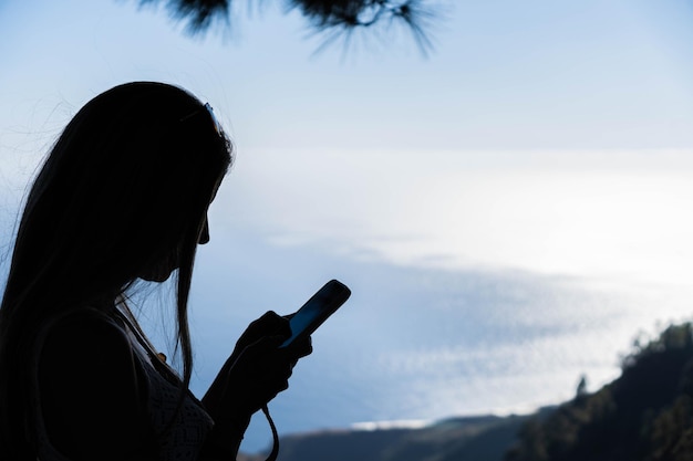 Silueta de mujer con cabello largo en la naturaleza escribiendo con su teléfono inteligente cerca del mar