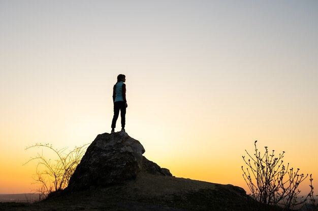 Silueta de una mujer caminante de pie solo en piedra grande al atardecer en las montañas.