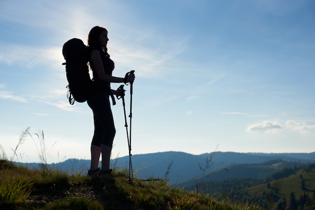 Silueta de mujer delgada turista con mochila y bastones de trekking de pie en la cima de una colina, mirando a otro lado contra el cielo azul de la mañana