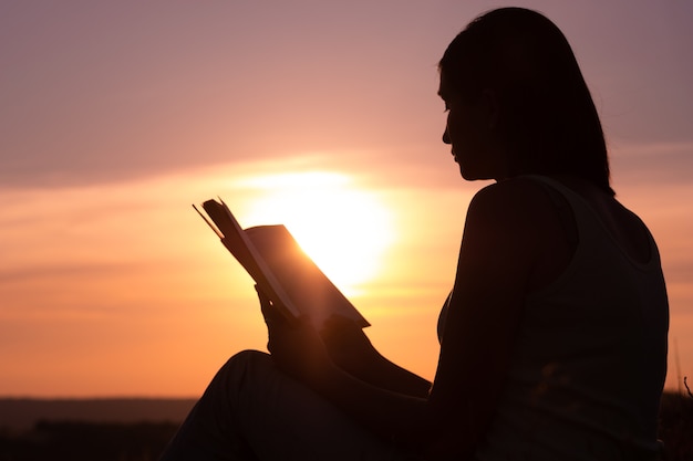 Silueta de una mujer hermosa joven al amanecer sentada en el suelo y mirando cuidadosamente el libro abierto