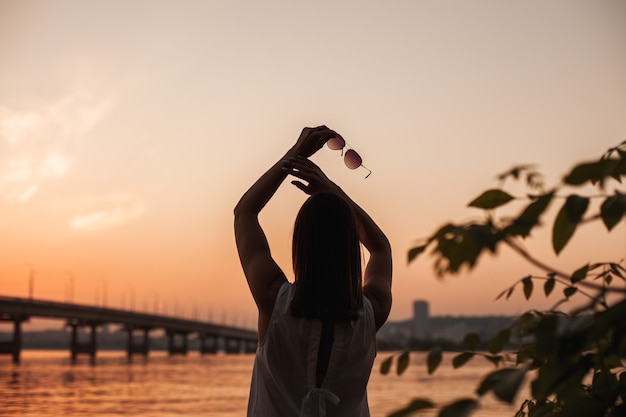 La silueta de una mujer joven en el cielo de verano al atardecer con un puente sobre el río y la silhou ...