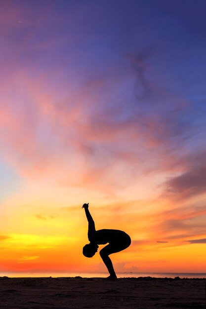 Silueta de mujer joven practicando yoga en la playa al atardecer.