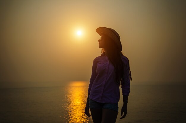 Silueta de mujer joven con un sombrero contra la puesta de sol sobre el mar. Hermosa chica delgada disfruta de paz y relajación en el océano