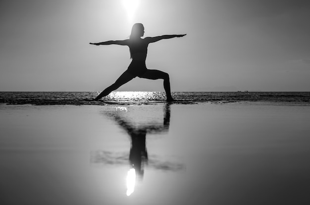 Silueta de mujer de pie en pose de yoga en la playa tropical durante la puesta de sol. Chica caucásica practicando yoga cerca del agua de mar. En blanco y negro