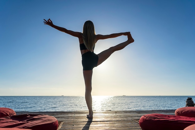 Foto silueta de mujer de pie en pose de yoga en la playa tropical durante la puesta de sol. chica caucásica practicando yoga cerca del agua de mar