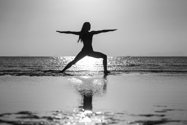 Silueta de mujer de pie en pose de yoga en la playa tropical durante la puesta de sol Chica practicando yoga cerca del agua de mar Blanco y negro