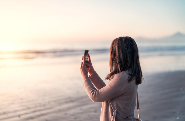 Silueta de una mujer en la playa al atardecer tomando fotos con un smartphone