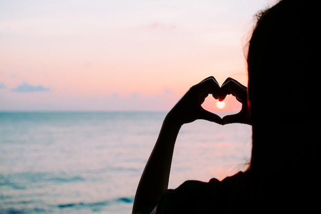 Silueta de mujer que hace el símbolo de forma de corazón por las manos con la puesta del sol y el océano dramáticos b