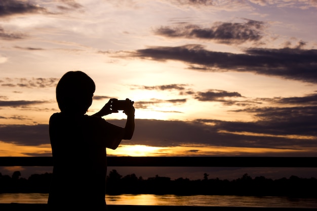 La silueta de la mujer que usa el teléfono elegante toma la foto sobre fondo hermoso de la puesta del sol.