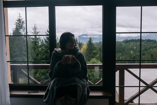 Silueta de una mujer sentada en el alféizar de la ventana con una taza