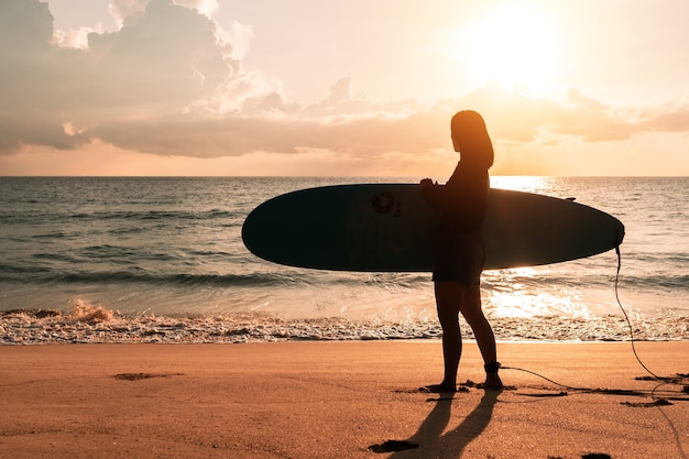 Silueta de mujer surfista llevando sus tablas de surf en la playa al atardecer con luz solar