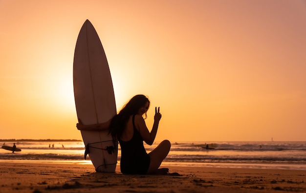 Foto silueta de una mujer surfista en una playa al atardecer haciendo el signo de la victoria